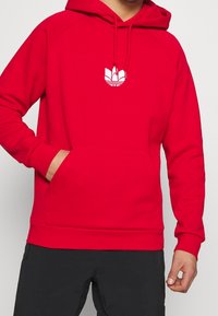 Rode hoodie van zacht materiaal, met een voorzak in kangaroo-stijl en een wit logo op de borst. Inclusief een capuchon met trekkoord.