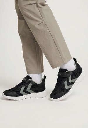 Piernas con pantalones beige claros y calcetines blancos con zapatillas negras de malla con rayas grises en forma de espiga y suelas blancas.