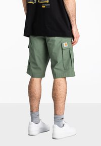 Pantalones cortos de carga verdes con múltiples bolsillos, hechos de tela duradera. Presenta un gran bolsillo lateral y una etiqueta de marca en el muslo derecho.