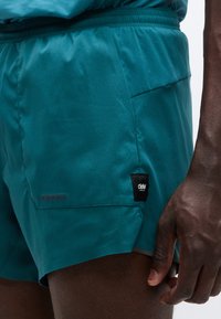 Τιρκουάζ αθλητικό σορτς από ελαφρύ ύφασμα, με πίσω τσέπη που έχει μαύρο λογότυπο και γκρι λεπτομέρειες στο τελείωμα.