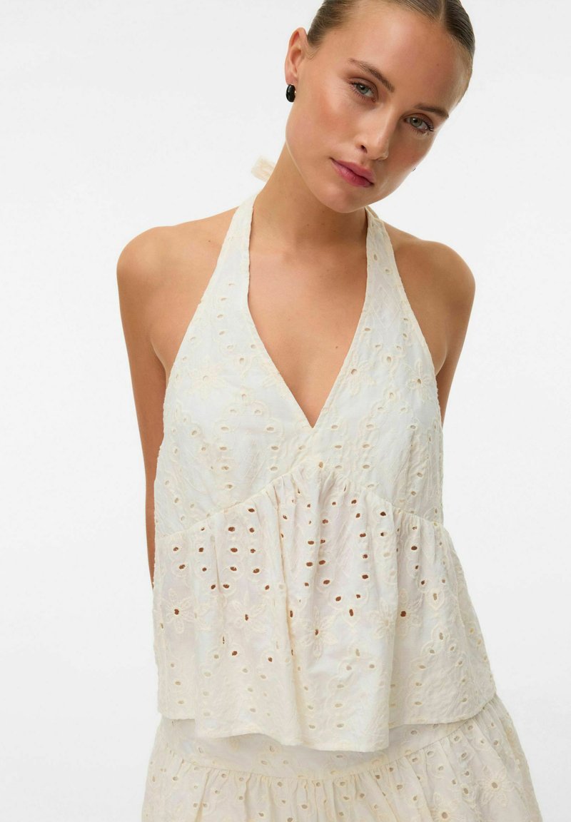 Jeune femme portant un haut dos nu en dentelle blanche avec un motif floral à œillets, debout devant un fond uni.