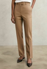 Femeie purtând pantaloni bej croiți cu pliuri frontale, pantofi slingback negri cu vârf ascuțit și un top alb băgat în pantaloni, stând pe un podea neutră.