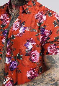 Camisa roja de manga corta con estampado floral en morado y rosa, llevada por una persona tatuada.