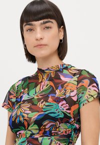 Jovem mulher com cabelo curto e escuro e brincos de argola, vestindo uma blusa transparente com um padrão tropical de folhas coloridas e laterais franzidas.