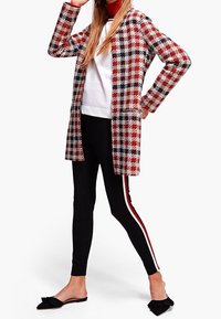 Kvinde iført en rød, sort og hvidternet frakke, hvid top, sorte leggings med røde og hvide side-striber, og sorte flade sko med sløjfe, står selvsikkert.