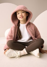 Vaaleanpunainen fleecetakki hupulla ja vetoketjulla, valkoinen t-paita, ruskeat ribbineulotut leggingsit ja valkoiset tennarit vaalein yksityiskohdin. Istuma-asento.