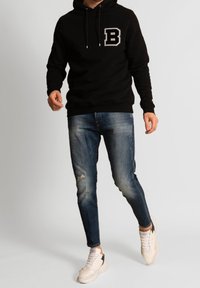 Musta huppari, jossa suuri valkoinen "B"-logo, yhdistetty kuluneisiin sinisiin farkkuihin ja vaaleanvärisiin tennareihin. Rentoon istuvuuteen ja kankaan tekstuuriin voi kiinnittää huomiota.