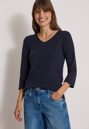 Femme souriante aux cheveux bruns portant un t-shirt bleu marine à manches longues avec col en V et un jean bleu, une main dans la poche.