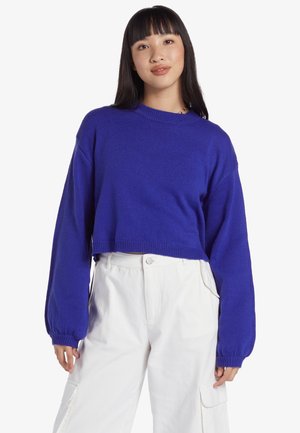 Gekürzter lila Pullover mit langen, ballonförmigen Ärmeln und gerippter Textur. Kombiniert mit weißen, hoch geschnittenen, weit geschnittenen Hosen.