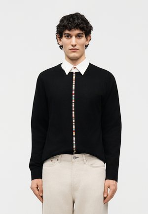 Νεαρός άνδρας με κοντά σγουρά μαλλιά, φοράει μαύρο πουλόβερ με λευκή γιακά και πολύχρωμες ρίγες, και ανοιχτό μπεζ παντελόνι.