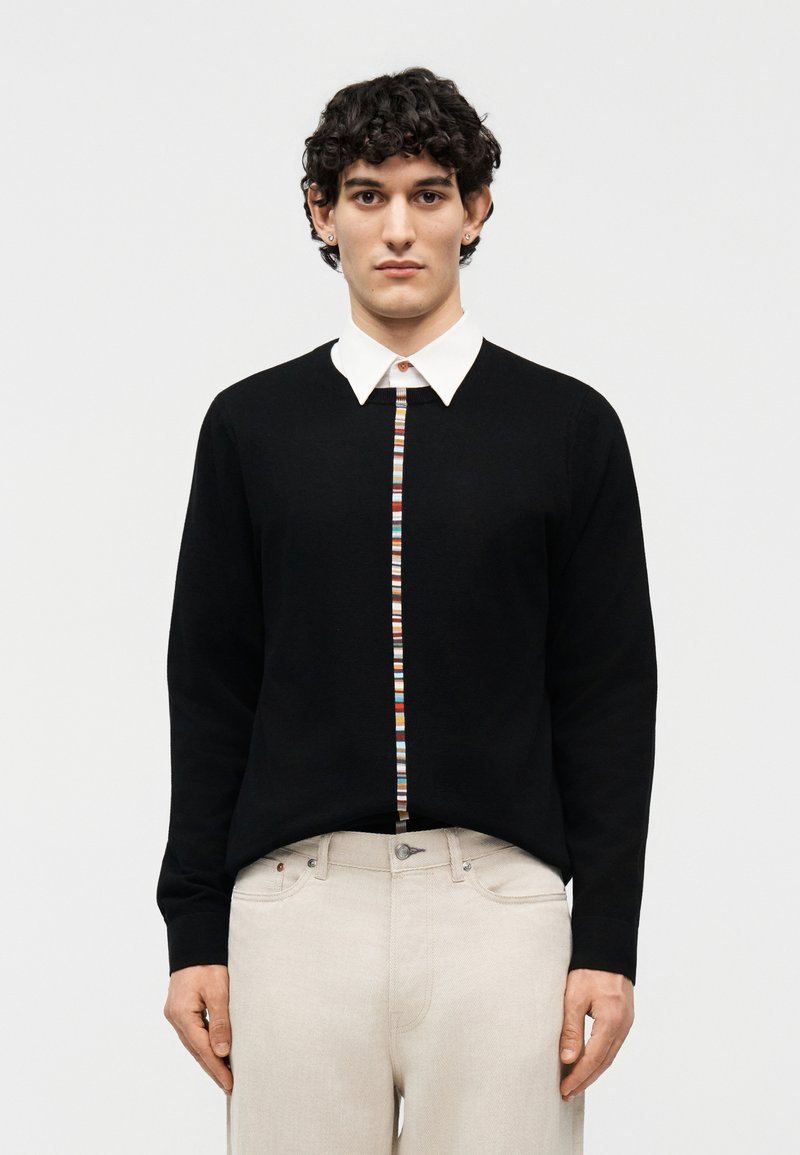 Νεαρός άνδρας με κοντά σγουρά μαλλιά, φοράει μαύρο πουλόβερ με λευκή γιακά και πολύχρωμες ρίγες, και ανοιχτό μπεζ παντελόνι.