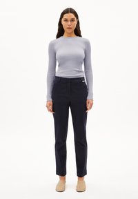 Lichtpaarse ribbed lange mouwen top gecombineerd met donkerblauwe op maat gemaakte broeken en beige instappers, staand voor een effen achtergrond.