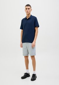 Marineblaues Poloshirt, kurze Ärmel, klassischer Kragen; kombiniert mit hellgrauen kurzen Hosen. Schwarze Schnürschuhe, weiße Crew-Socken, sauberer Hintergrund.