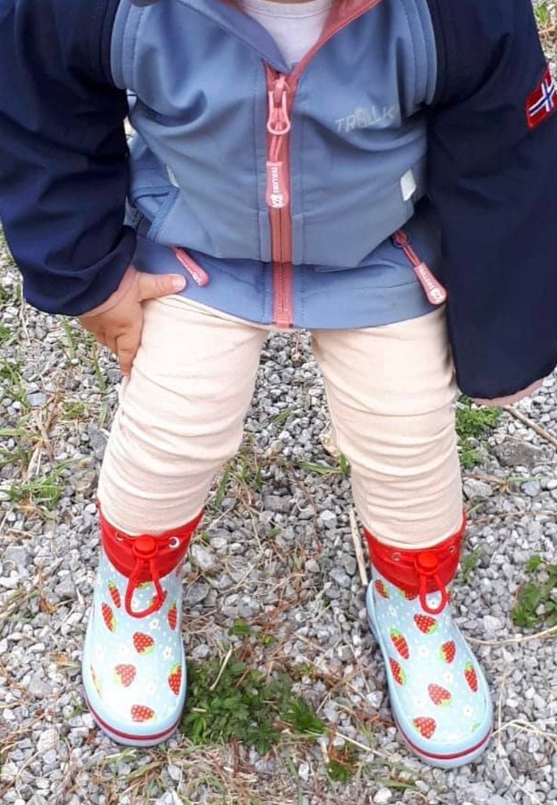 Bottes en caoutchouc colorées avec un fond bleu clair, des accents rouges et un motif de fraises. Portées par-dessus des pantalons beige clair.