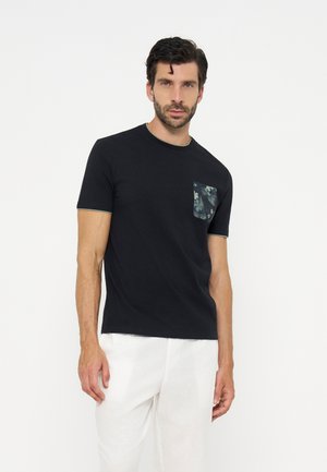 Homme aux cheveux bouclés foncés portant un t-shirt noir à manches courtes avec une poche poitrine à motif et un pantalon blanc, debout sur un fond blanc.