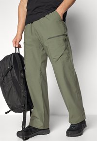 Olive green cargo pants with multiple pockets, including a zippered one. Made from lightweight material. Worn with a black backpack and shoes.