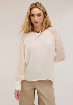 Mujer con cabello largo y rubio que lleva una blusa blanca transparente de manga larga y pantalones beige, de pie frente a un fondo liso.