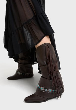 Sötétbarna, térdig érő bőrcsizma rojttal és türkiz berakásos ezüst koncsókkal, áttetsző fekete szoknya alatt viselve.