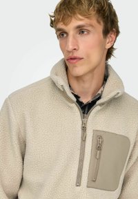 Kermansävyinen fleece-pulloveri, jossa on korkea kaulus, etuvetoketju ja teksturoitu pinta. Vasemmassa rinnassa on vaaleanruskea vetoketjullinen tasku.