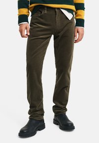 Oliwkowe spodnie z materiału corduroy o slim fit, z zamkiem błyskawicznym, pięcioma kieszeniami i subtelną fakturą. Połączone z czarnymi skórzanymi butami.