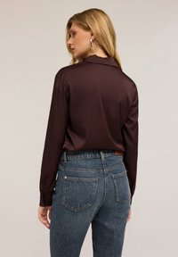 Vrouw met lang blond haar, gekleed in gouden oorbellen, een donkerbruine satijnen blouse en hoge blauwe jeans, met haar rug naar de camera gekeerd tegen een effen achtergrond.