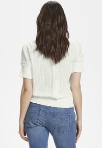 Blusa bianca a trama con maniche corte, con strisce verticali e inserti in pizzo lungo il retro e le maniche. Abbinata a jeans blu.