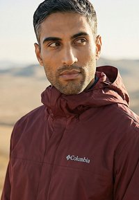 Man draagt een bordeauxrode Columbia-jas met capuchon en kijkt opzij in een buitenomgeving met een wazige natuurlijke achtergrond.