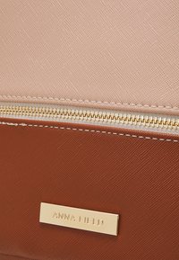 Gros plan d'un portefeuille beige et marron texturé avec une fermeture éclair dorée et une plaque métallique dorée portant le logo « ANNA FIELD ».