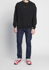 Persona che indossa una felpa nera con un piccolo logo, jeans blu scuro e scarpe bianche con motivi rossi e neri, in piedi su uno sfondo neutro.