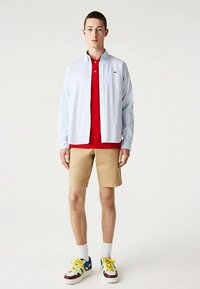 Gaiši zila krekls ar garām piedurknēm virs sarkanas polo krekla, bēšas šorti un daudzkrāsainas sporta kurpes. Krekls ir ar pogām un logo.