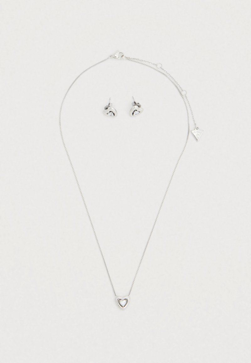 Σετ ασημένια κοσμήματα που περιλαμβάνει ένα λεπτό μενταγιόν σε σχήμα καρδιάς σε αλυσίδα και τα αντίστοιχα σκουλαρίκια καρφάκια με αντανακλαστικές λεπτομέρειες.