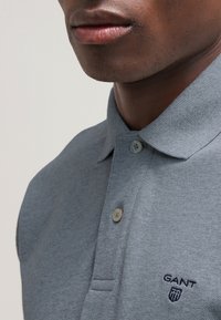Gros plan d'une personne portant un polo gris avec deux boutons et un petit logo GANT sur la poitrine.