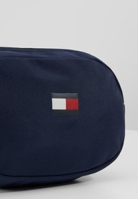 Granatowa torba z teksturowanej tkaniny, z małym prostokątnym logo w kolorach czerwonym, białym i czarnym. Zamek błyskawiczny wzdłuż górnej krawędzi.
