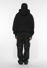 Sudadera negra de gran tamaño con capucha y cordón, combinada con pantalones de cuero negros de corte holgado, vista desde la parte trasera sobre un fondo blanco.