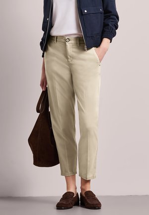 Osoba ubrana w beżowe spodnie 7/8, biały top, granatową kurtkę, brązowe mokasyny, trzyma dużą brązową torbę zamszową wzdłuż ciała.