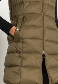 Olivgrüne Stepp-Weste mit horizontalen Nähten, einem mittigen Reißverschluss und einer Tasche mit Druckknopf. Der Stoff ist glatt mit leichtem Glanz.