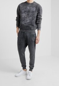 Person med en mörkgrå acid-wash sweatshirt och joggers med dragkedjefickor, kombinerat med vita sneakers, som står framför en enkel bakgrund.