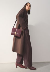 Manteau en laine marron sur un pantalon bordeaux profond avec un sac à main assorti en cuir embossé croco. Des bottes à talons bordeaux brillantes complètent l'ensemble.