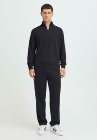 Un homme debout portant un sweat-shirt noir zippé et un pantalon noir avec des baskets blanches à rayures noires sur un fond clair.