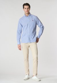 Camicia button-down a righe blu chiaro e bianche con colletto, abbinata a pantaloni beige e sneakers bianche, in piedi contro uno sfondo neutro.