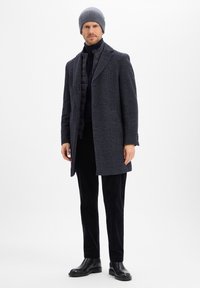 Marineblauer Tweed-Mantel mit strukturiertem Muster, über einem schwarzen Rollkragenpullover und einer Weste getragen, kombiniert mit dunklen Cordhosen und schwarzen Stiefeln.