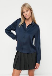 Navy blue satin shirt with a collared neckline and button front, paired with a pleated dark gray skirt. Smooth texture and relaxed fit.