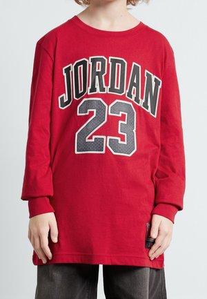 Barn iført en rød langærmet trøje med "JORDAN 23" trykt i sort og hvid på fronten, kombineret med mørke bukser.