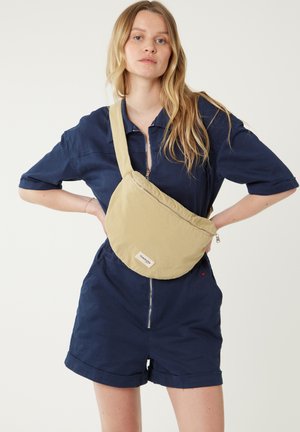 Sac en toile jaune de forme arrondie, avec fermeture éclair et petite étiquette de marque. Porté sur une combinaison bleu marine.