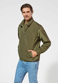 Lekka oliwkowa kurtka z wysokim kołnierzem, zamkiem z przodu i bocznymi kieszeniami, w połączeniu z niebieskimi dżinsami. Gładka, błyszcząca tekstura.