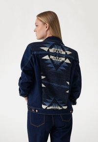 Džinsinis švarkas su tamsiai mėlynu korpusu ir raštuotu trikotažo audiniu ant nugaros, kuriame yra geometriniai raštai mėlynos ir kreminės spalvose.