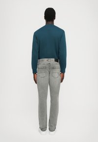 Mörk teal sweatshirt med långa ärmar, kombinerad med ljusgrå jeans. Jeansen har bakfickor och en svart etikett på midjebandet.