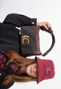 Una donna tiene una borsa bordeaux con un dettaglio di leopardo dorato, indossando un cappello a secchiello bordeaux e una giacca nera, mostrando un outfit elegante.