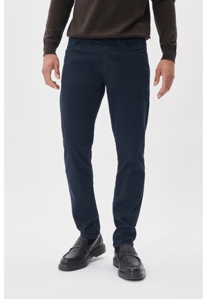 Mies, jolla on tummansininen slim fit -housut, mustat loaferit ja ruskea pitkähihainen yläosa, seisoo yksinkertaista valkoista taustaa vasten.