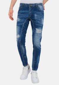 Blauwe spijkerbroeken met verweerde details, lichte spetterpatronen en een slim fit. Gecombineerd met witte sneakers.